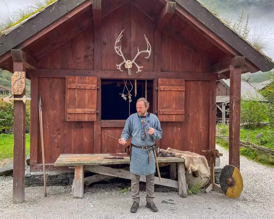 A man stands in front of a wooden building with a grass roof, holding a stick.