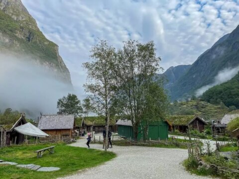 A wide shot of a Viking village with wooden buildings and grass roofs, nestled in a valley with mountains on either side.
